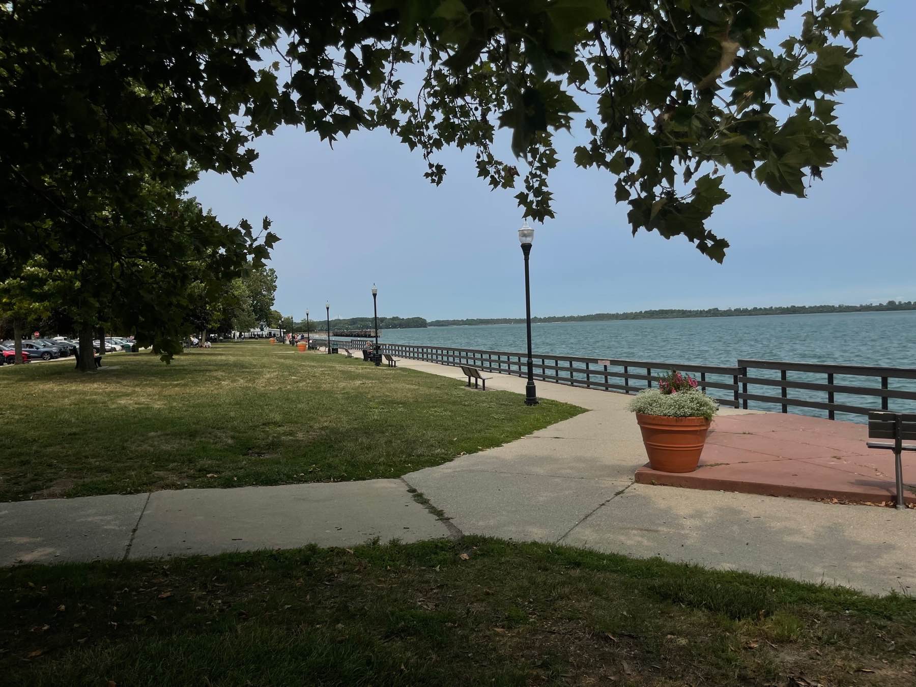 Bishop Park along the Detroit River in Wyandotte — a tree-lined waterfront promenade with benches, a lamp post, and a flowering planter on a soft, overcast day